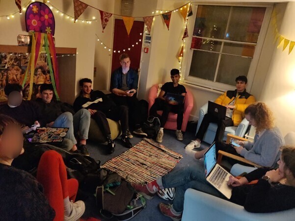 A group of friends in a room with bunting, fairy lights and a rug, using laptops.